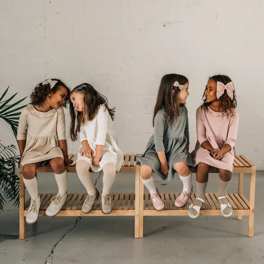Four young girls sit on a wooden bench, each wearing pastel-colored dresses and knee-high socks paired with Zimmerman Shoes’ Mini Boys Saddles in Sand & Fog. Two girls chat on the left while the other two share smiles on the right, all set against a light background.