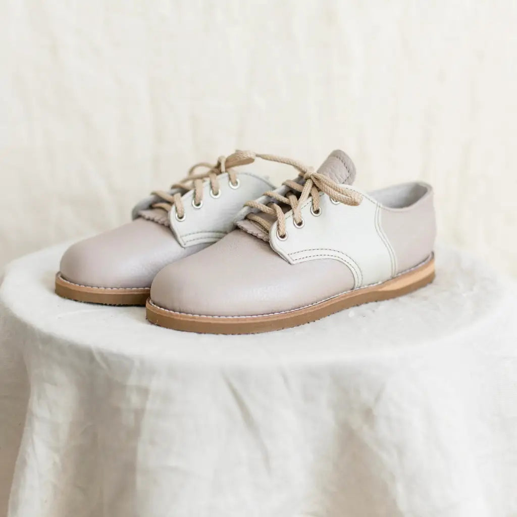 A pair of Zimmerman Shoes Mini Boys Saddles in Sand & Fog, featuring tan laces, sits on a round, white, textured pedestal against a neutral background.