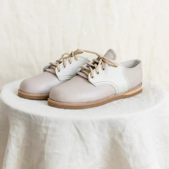 A pair of Zimmerman Shoes Mini Boys Saddles in Sand & Fog, featuring tan laces, sits on a round, white, textured pedestal against a neutral background.