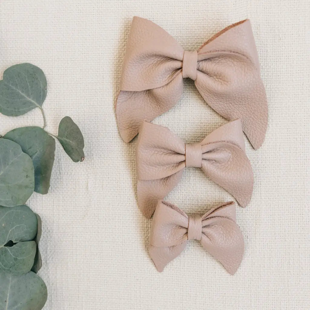 Three beige leather Mini Boys Saddles in Sand & Fog by Zimmerman Shoes, shown in small, medium, and large sizes, are arranged vertically on a textured cream fabric background—evoking the refined details of formal footwear. Green eucalyptus leaves are placed to the left of the shoes.