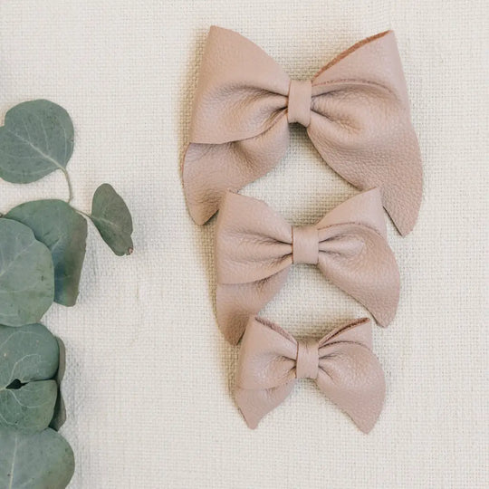 Three beige leather Mini Boys Saddles in Sand & Fog by Zimmerman Shoes, shown in small, medium, and large sizes, are arranged vertically on a textured cream fabric background—evoking the refined details of formal footwear. Green eucalyptus leaves are placed to the left of the shoes.