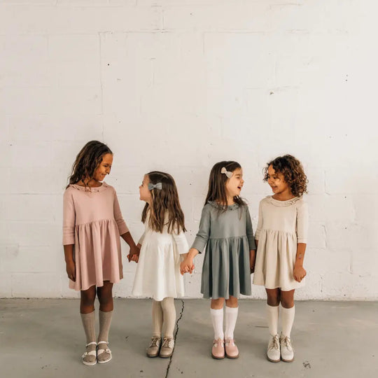 Four young girls in pastel dresses and knee-high socks stand in a row, holding hands and smiling at each other against a plain white wall, their outfits completed with Zimmerman Shoes’ Mini Boys Saddles in Sand & Fog for a playful, polished touch.