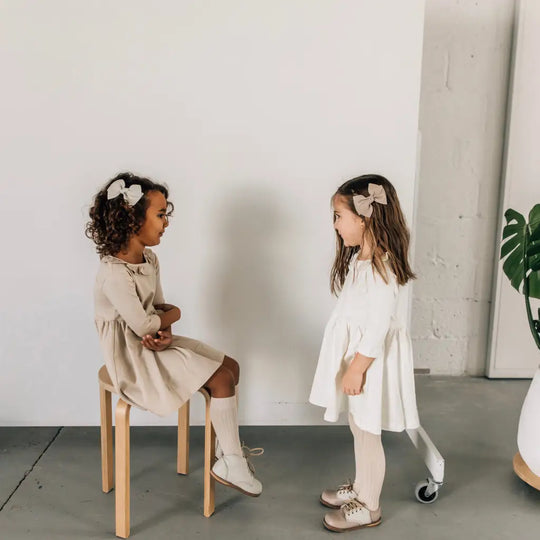 Two young girls in neutral-colored dresses and hair bows face each other indoors. One, seated on a wooden stool, wears the Zimmerman Shoes Mini Boys Saddles in Sand & Fog, while the other stands nearby. A green plant is partially visible to the right in the minimalist setting.