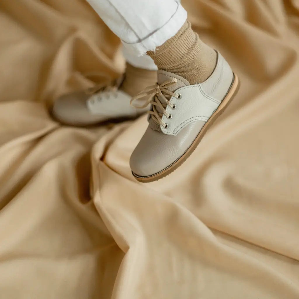 A person wearing Zimmerman Shoes’ Mini Boys Saddles in Sand & Fog stands on a light tan fabric backdrop, paired with beige socks and white pants.