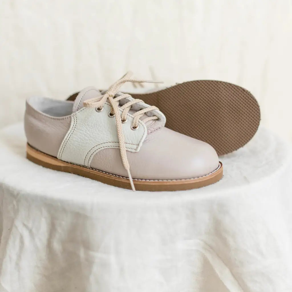 A pair of Zimmerman Shoes Mini Boys Saddles in Sand & Fog, crafted with beige leather and off-white panels, rests atop a round, white fabric-covered surface. One shoe stands upright while the other leans on its side to reveal the brown textured sole—emphasizing their refined style as formal footwear.