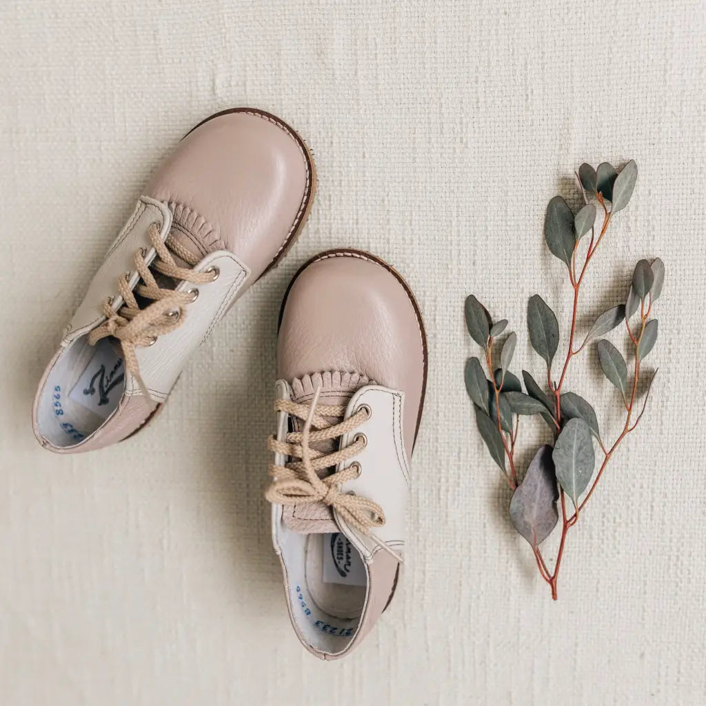 A pair of Zimmerman Shoes Mini Boys Saddles in Sand & Fog, crafted from soft beige leather with taupe lace-up details, displayed beside a eucalyptus branch on a textured, light-colored surface.