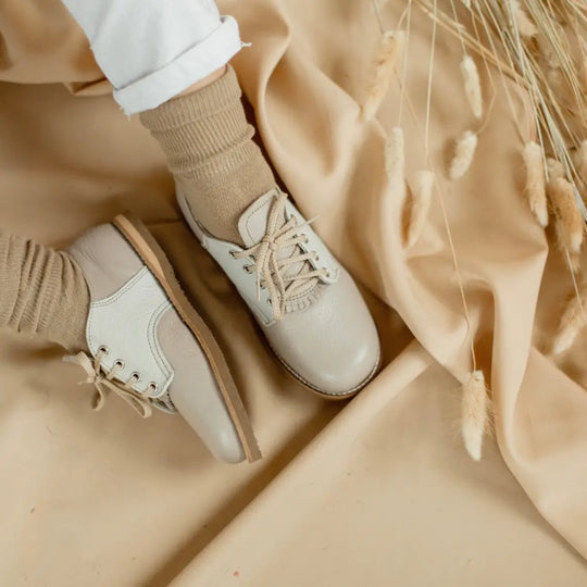 Wearing Zimmerman Shoes’ Mini Boys Saddles in Sand & Fog, a person with beige socks sits on a beige fabric adorned with dried grass decorations.