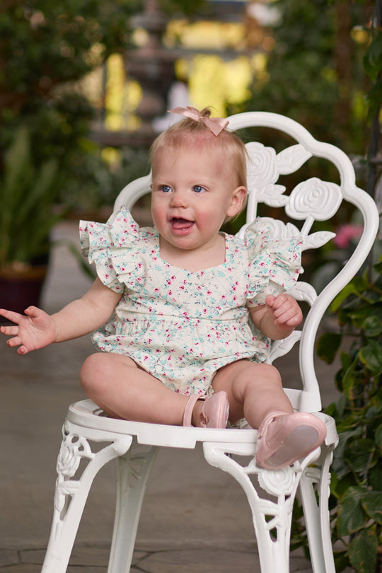 A smiling baby with blonde hair and a pink bow sits on a white, ornate chair, wearing the charming Baby Hattie Romper in Pansy Trellis by Ivy City Co—a floral romper paired with pink shoes—surrounded by greenery.