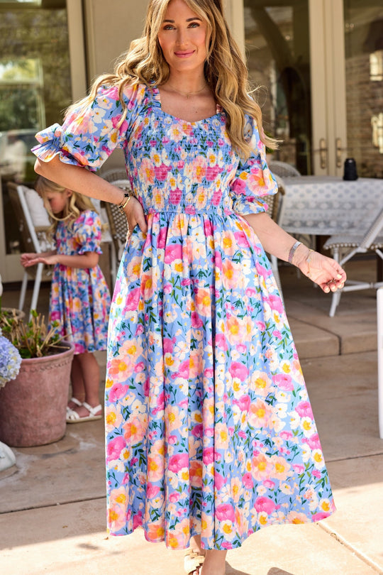 A woman in a colorful floral midi dress stands outside, smiling. Nearby, a young girl with long hair wears the Ivy City Co Teagan Dress in Delia Blue and stands near a table and chairs on the patio amid outdoor decor.