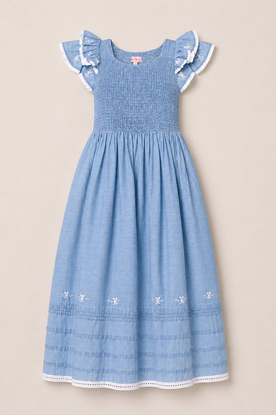 The Hattie Ankle Length Dress in Embroidered Chambray by Ivy City Co is a light blue, sleeveless dress with a smocked bodice for a comfortable and maternity-friendly fit. It features ruffled cap sleeves with white trim and embroidered floral details near the hem, and is displayed on a neutral background.