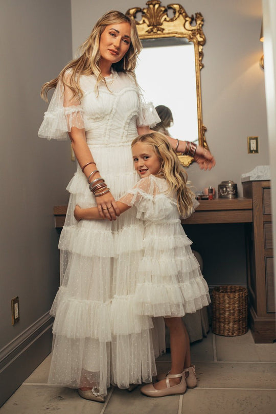 A woman and a young girl, both wearing the Ivy City Co Whimsical Dress in White, stand together in front of a vanity mirror. Smiling as the girl hugs the woman’s waist, their matching dresses glow softly in the warm lighting of the beautifully decorated room.