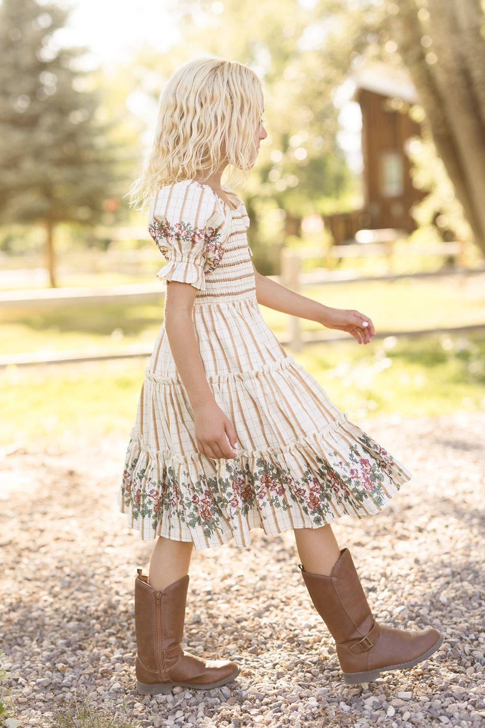 A young child with blonde hair walks along a gravel path outside, sunlight streaming through the trees, wearing the Ivy City Co Mini Madeline Dress in Embroidered Cider Grid, which features a smocked bodice and charming puff sleeves.