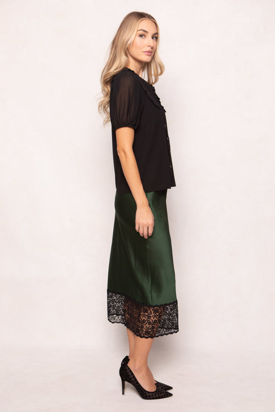 A woman with long, wavy blonde hair stands in profile against a plain light background, wearing the Andrea Blouse in Black Chiffon by Ivy City Co. She pairs it with a green satin skirt featuring black lace trim and black high heels.
