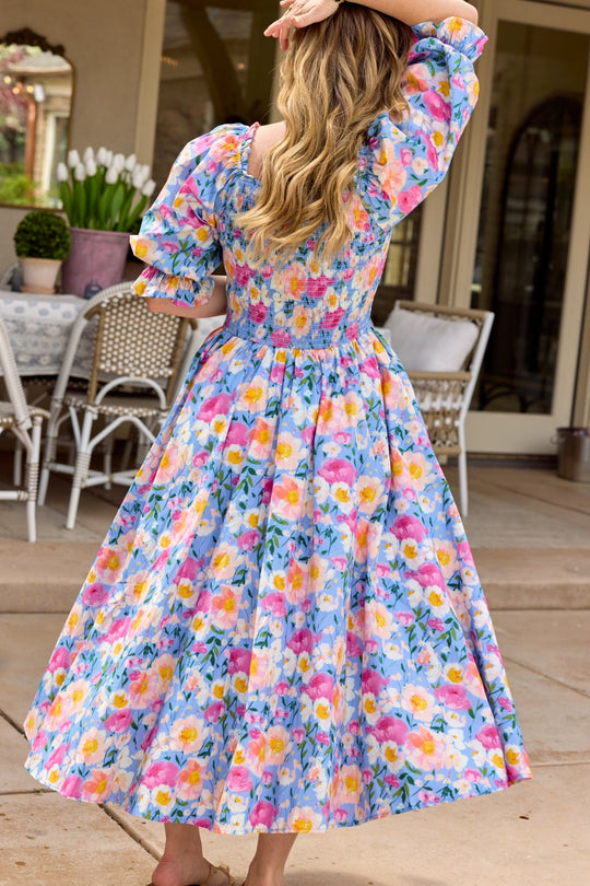 A woman with long, wavy blonde hair wears the Teagan Dress in Delia Blue by Ivy City Co, featuring a colorful floral pattern. She stands barefoot outside with patio furniture and potted tulips visible in the background, facing away from the camera.