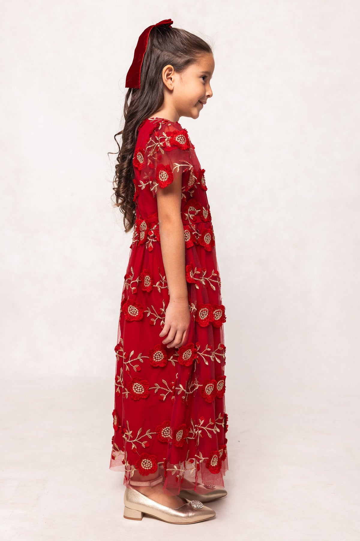 A young girl stands in profile, wearing the Ivy City Co Mini Athena Dress in Red—an elegant girls’ maxi dress adorned with delicate floral embroidery. She pairs it with gold shoes, her long wavy hair tied back with a red ribbon, and smiles gently while posed against a simple light backdrop. This beautiful dress is perfect for holidays or special mommy and me moments.