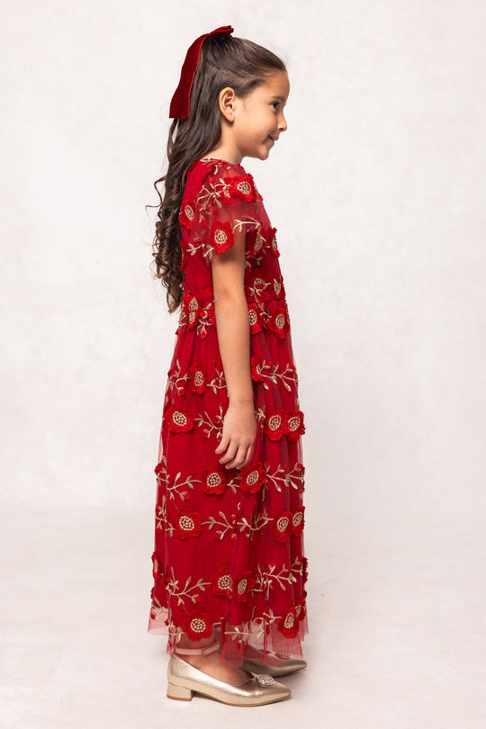A young girl stands in profile, wearing the Ivy City Co Mini Athena Dress in Red—an elegant girls’ maxi dress adorned with delicate floral embroidery. She pairs it with gold shoes, her long wavy hair tied back with a red ribbon, and smiles gently while posed against a simple light backdrop. This beautiful dress is perfect for holidays or special mommy and me moments.