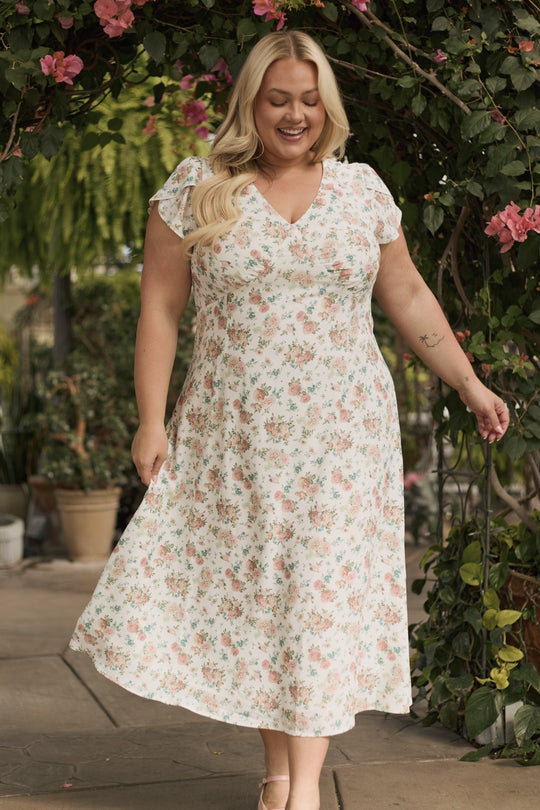 A woman wearing the Fiona Dress in Countryside Peony Blossom by Ivy City Co smiles as she walks outdoors beneath a leafy archway adorned with pink flowers.