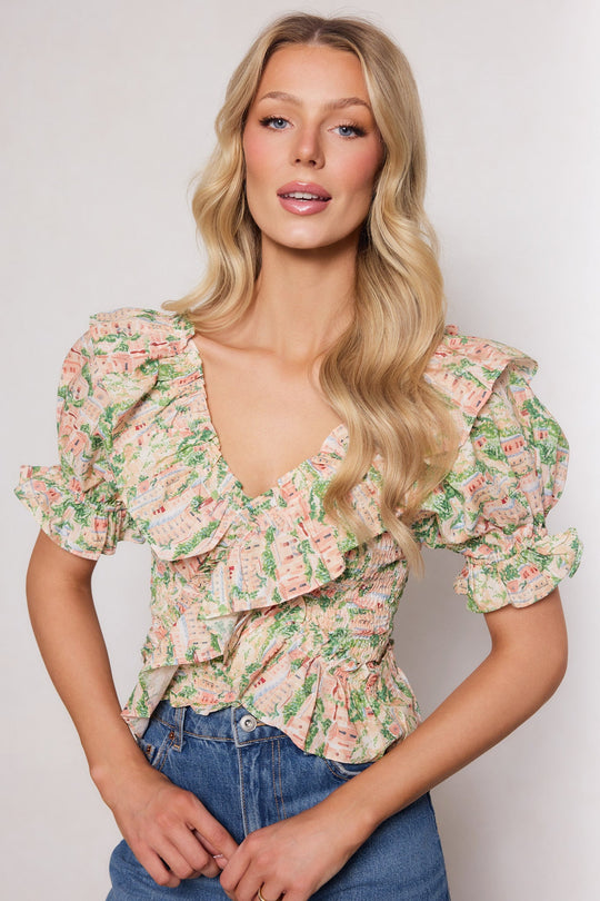 A woman with long blonde hair stands against a plain light background, facing forward and smiling slightly, wearing the Ivy City Co Julian Top in Cream—a floral ruffled blouse with puffed sleeves—paired with blue jeans.