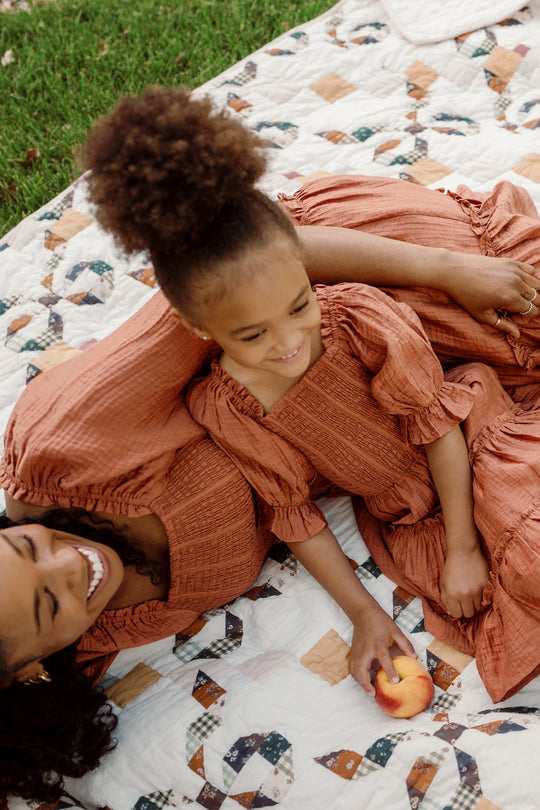A woman and a young girl, both in matching rust-colored dresses with puff sleeves, smile and relax on a patterned quilt outdoors. The girl, dressed in Ivy City Co’s Mini Madeline Dress in Apple Cider with its charming smocked bodice, holds a peach while leaning against the woman.
