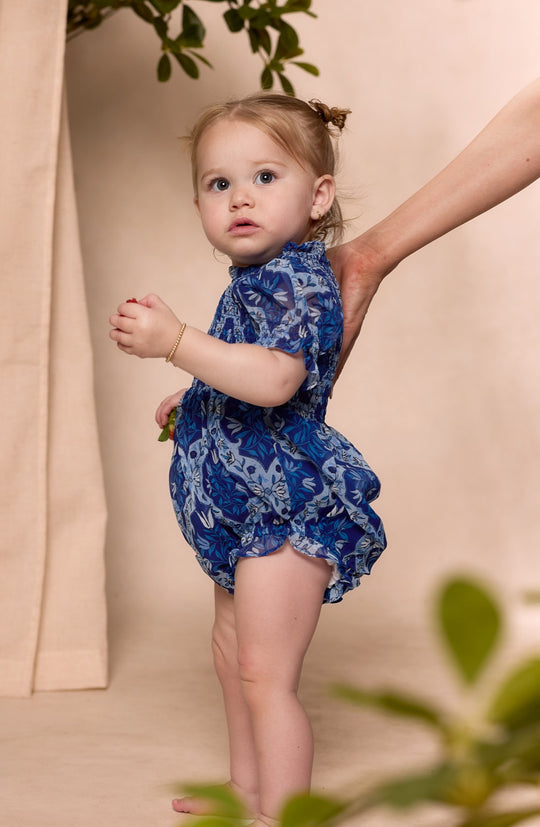 A young child with light hair wears the Ivy City Co Baby Cordelia Romper in Trellis Foulard—a blue floral smocked romper. The child stands sideways, looking up as an adult hand supports their back, with green leaves and a beige curtain behind.
