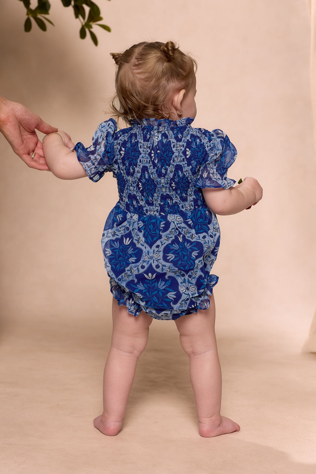 A toddler wears the Ivy City Co Baby Cordelia Romper in Trellis Foulard, featuring a smocked bodice and blue patterned fabric, standing on a beige surface while an adult hand gently supports their arm.