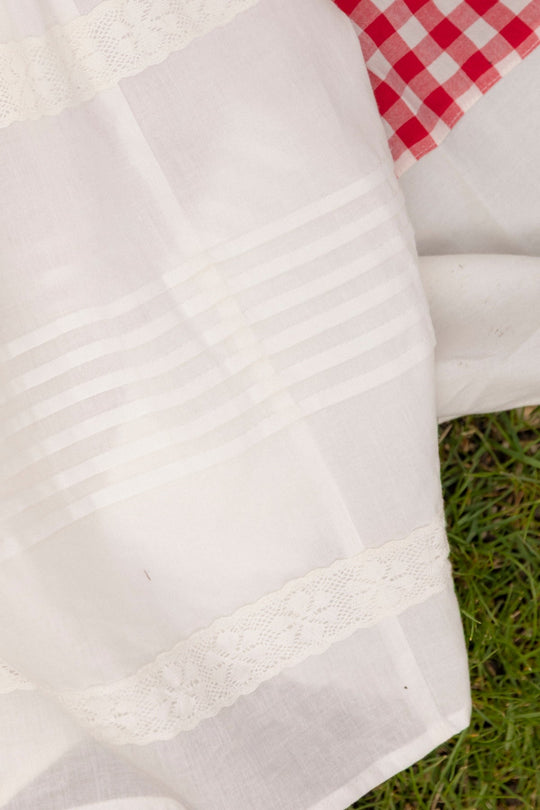 A close-up of the Ivy City Co Mini Palermo Dress in White showcases its white cotton fabric with lace details and horizontal stripes, resembling a smocked bodice with delicate puff sleeves, placed beside a red and white checkered cloth on the grass.