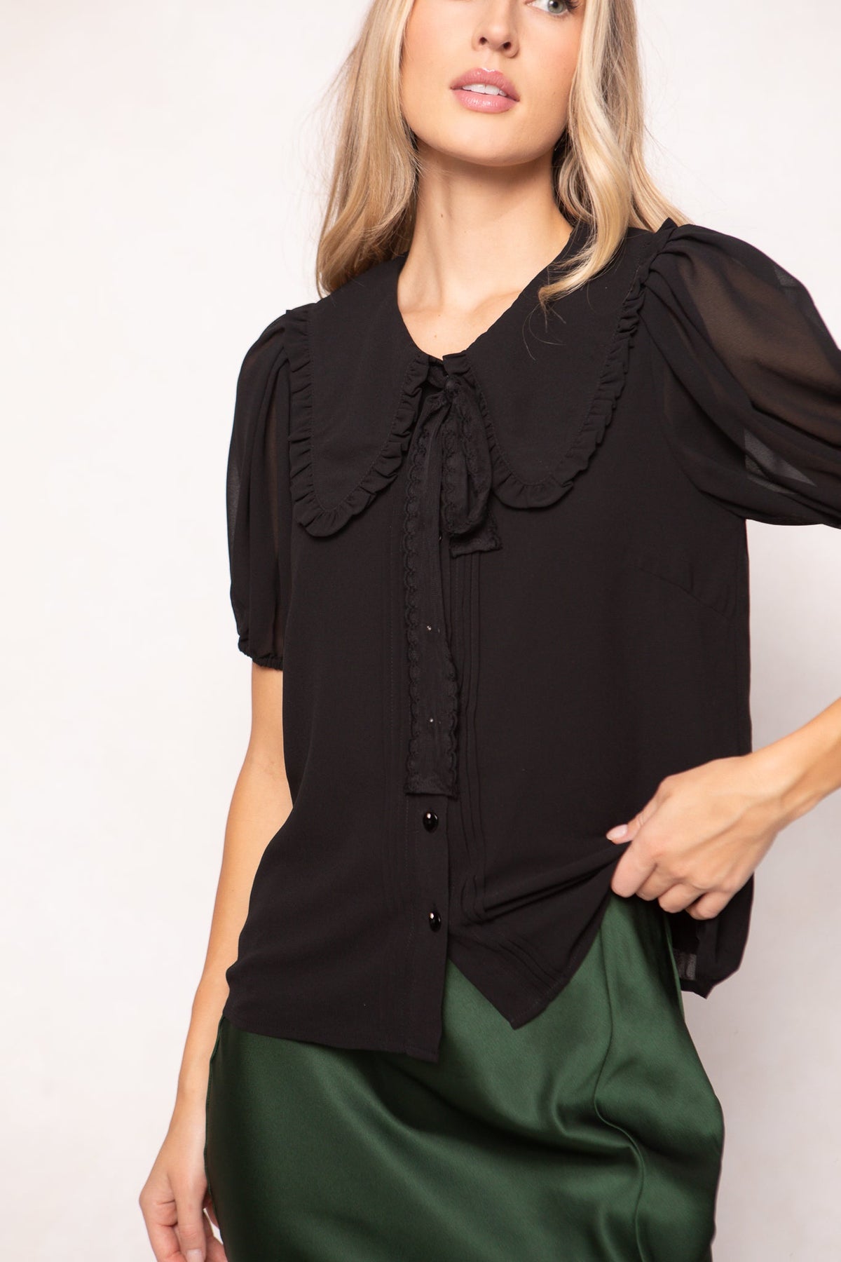 Dressed in the Andrea Blouse in Black Chiffon by Ivy City Co, a woman poses elegantly with one hand on her hip against a light background. The blouse features striking, oversized ruffles and a bow at the collar, perfectly paired with a shiny green skirt.