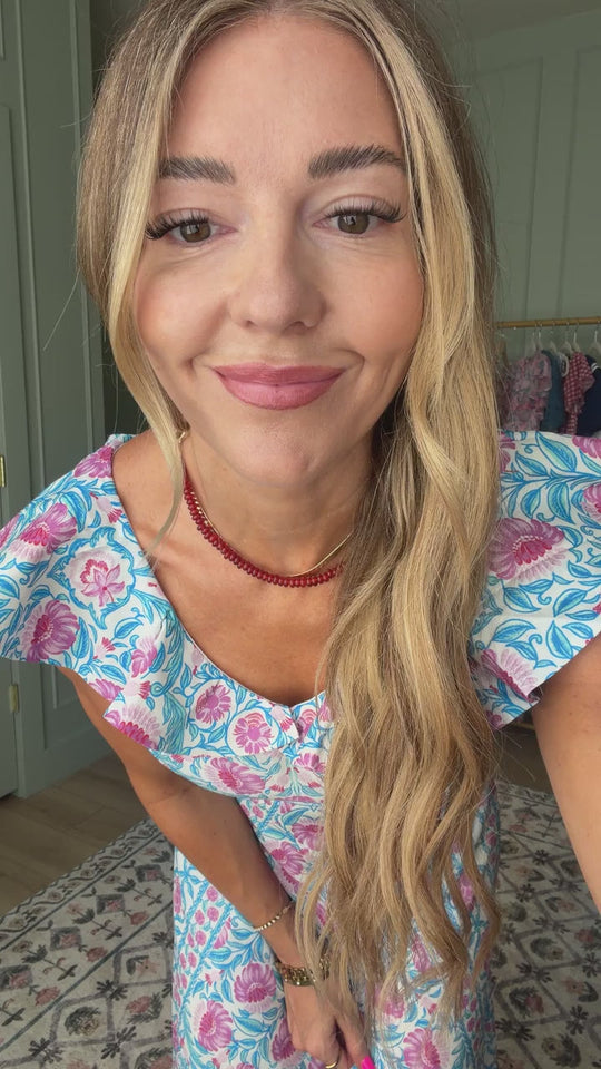 A woman with long blonde hair, wearing the Ivy City Co Samara Dress in Thistle Flower Mosaic Florise and a red necklace, smiles at the camera in a room with patterned carpet and clothing hanging in the background.