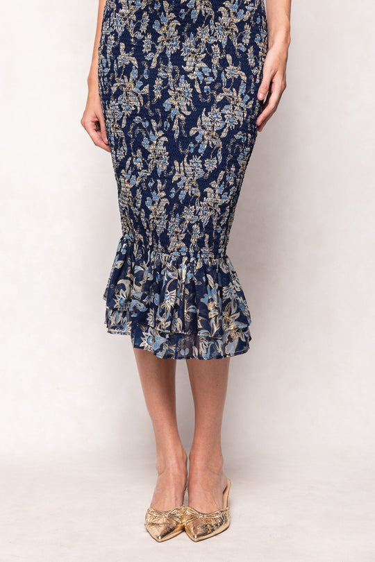 A person wearing the Mira Dress in Gilded Vine by Ivy City Co—featuring a dark blue floral pattern and a ruffled hem—paired with beige pointed-toe shoes, shown from mid-torso down against a light background.