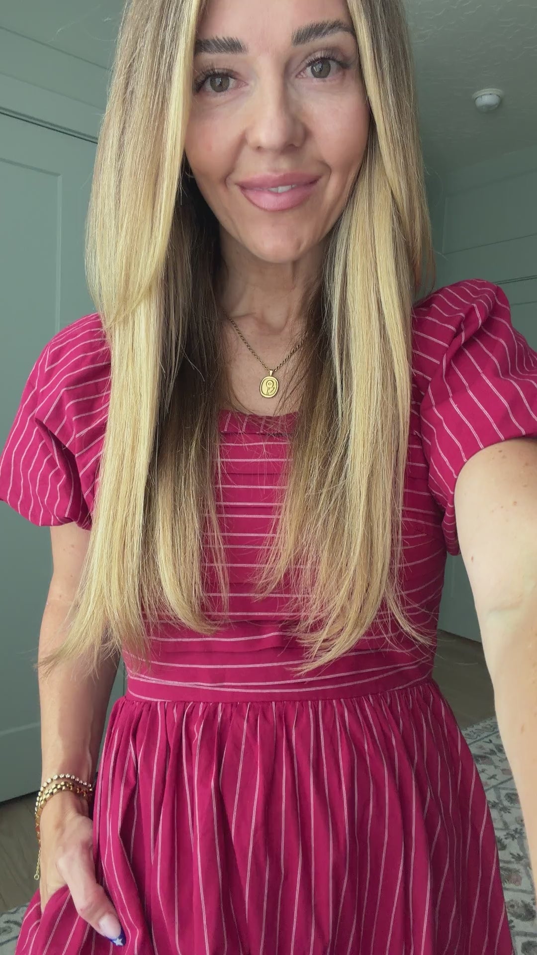 A smiling woman with long blonde hair wears the Ivy City Co Julie Dress in Cranberry Pinstripe, featuring a smocked bodice and puff sleeves, standing indoors.