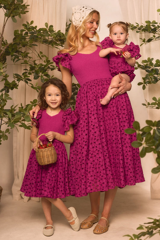 Three smiles shine among green plants as a woman and two young girls wear matching Ivy City Co Mini Hattie Dresses in magenta, featuring beautiful embroidered eyelet floral patterns. The woman cradles the baby while the older girl, dressed in a girls’ cotton midi version of the Mini Hattie Dress, holds a small basket—capturing a perfect Mini & Me Easter moment.