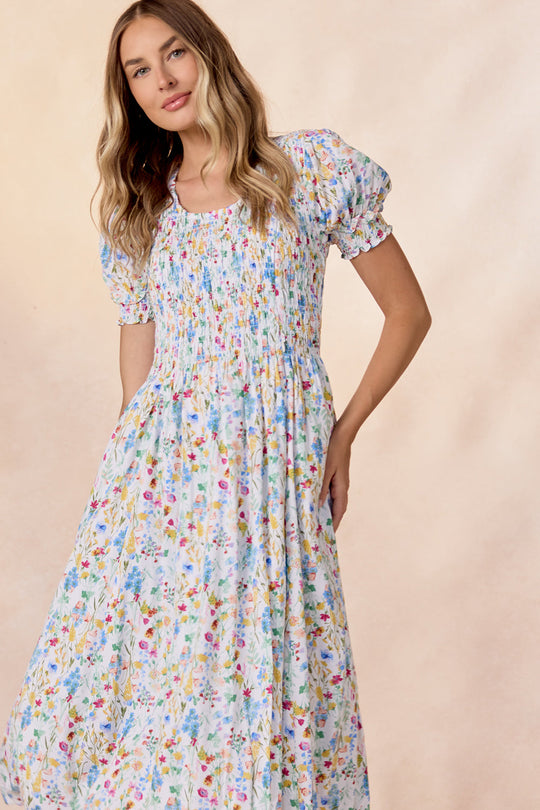 A woman wearing the Amelie Dress in Floral Fields by Ivy City Co—a light-colored, short-sleeve floral midi dress featuring a shirred bodice and puffed sleeves—stands against a soft beige background. This charming maternity dress offers both style and comfort for expecting mothers.