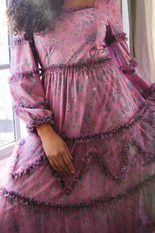 A person wearing the Ivy City Co Catherine Dress in Climbing Vine Bloom—a long-sleeved, ruffled, floral pink dress—stands near a window with one hand resting on the skirt. Their face is not visible.