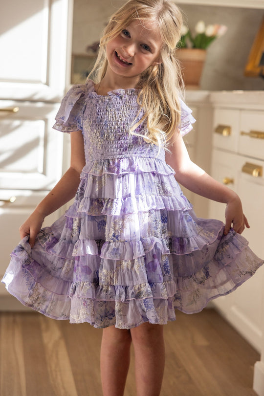 A smiling young girl poses playfully in a bright kitchen, holding the sides of her Ivy City Co Mini Winnie Dress in Lavender Aquarelle Fleur, featuring a ruffle tiered skirt and smocked bodice.