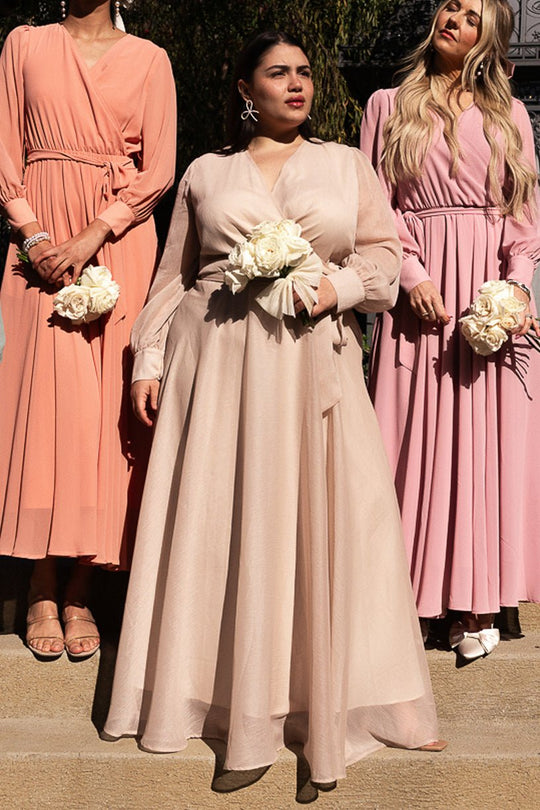 Three women wearing the Ivy City Co Andie Dress in Champagne stand side by side outdoors, each holding a bouquet of white roses. Their expressions are neutral, and sunlight casts gentle shadows on their champagne-colored dresses.