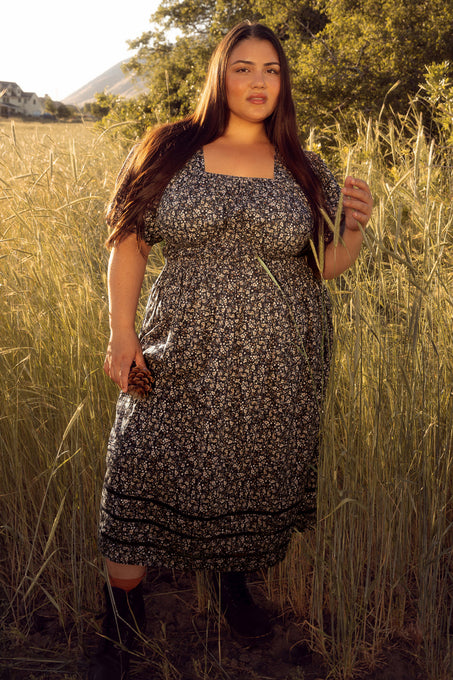 Woman in a floral dress standing in a field with tall grass and trees in the background.