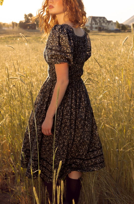 Woman in a floral dress standing in a field with a sunset or sunrise in the background