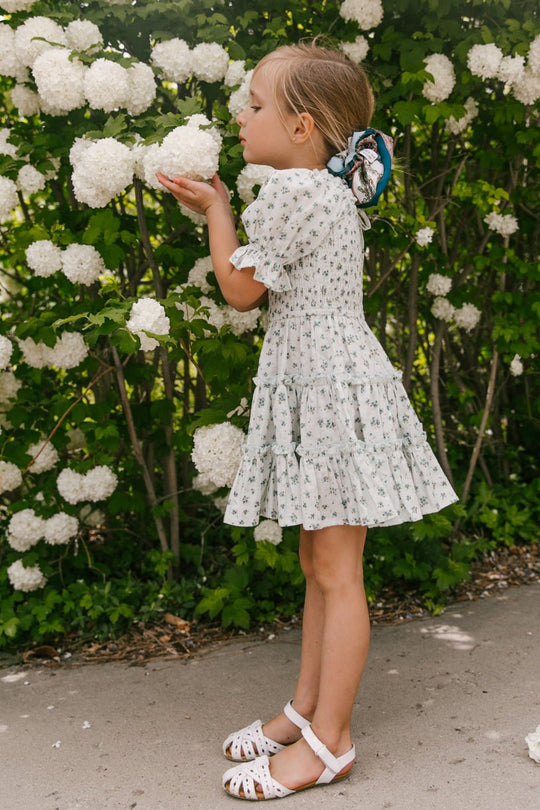 A young girl wearing the Ivy City Co Mini Madeline Dress in Blue and sandals smells a cluster of white flowers on a bush, surrounded by lush greenery and blooms in the background.