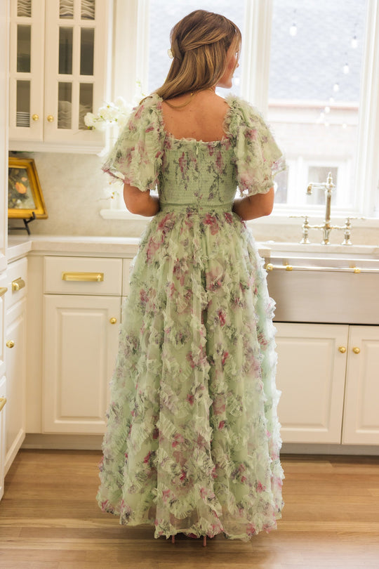 A woman with long hair stands in a sunlit kitchen, her back to the camera. She wears Ivy City Co’s Florentina Dress in Pistachio Aquarelle Fleur—a flowing chiffon design with ruffled sleeves that’s both maternity and nursing friendly—as she gazes out a window above a farmhouse sink.