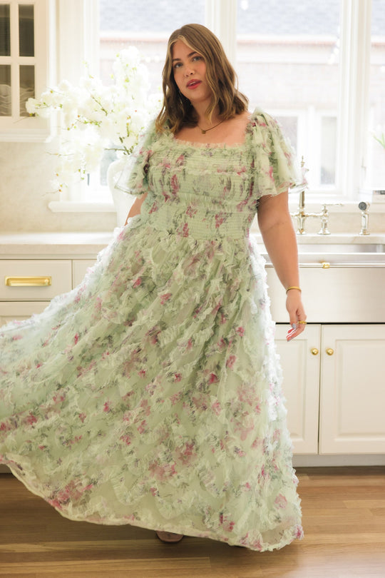 A woman poses in the Ivy City Co Florentina Dress in Pistachio Aquarelle Fleur, a nursing-friendly and maternity-friendly chiffon dress, in a bright kitchen with white cabinets, a large window, and flowers on the counter.
