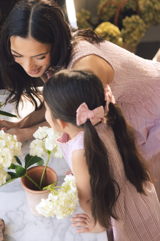 A woman and a young girl with long hair, both dressed in matching pink outfits from Ivy City Co, admire white hydrangeas on a marble table. The woman’s effortlessly elegant look is thanks to the smocked Karla Dress in Pink Striped Gingham, while the girl accessorizes her outfit with a coordinating pink bow.