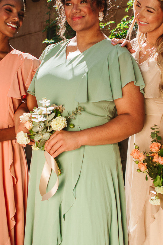 Three women in pastel dresses stand together, smiling. The woman in the center wears the Ivy City Co Callie Dress in Sage Chiffon and holds a bouquet of white flowers. Sunlight illuminates their faces and floral bouquets.