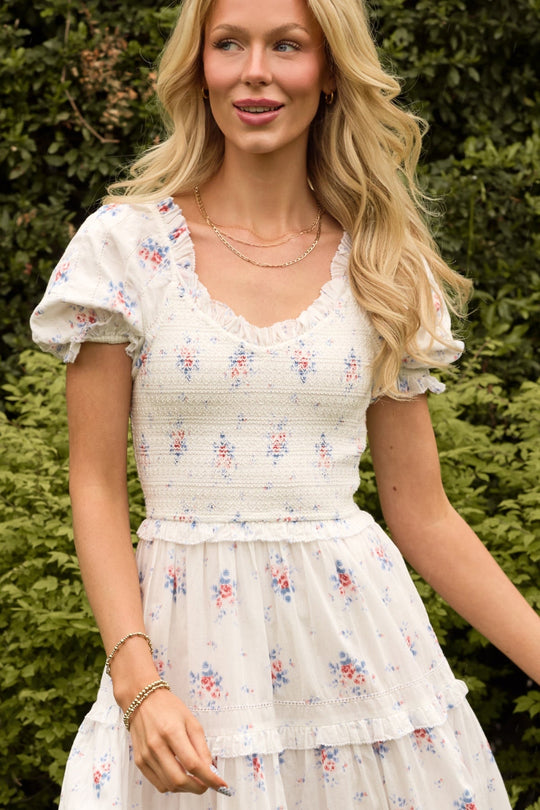 Wearing the Emily Dress in Heritage Soft Ditsy Floral by Ivy City Co—a white, nursing-friendly midi dress with puff sleeves—a woman stands outdoors in front of green bushes, smiling and looking to the side.