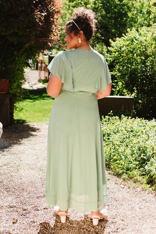 A woman with curly hair stands on a gravel path amid lush greenery, wearing the Ivy City Co Callie Dress in Sage Chiffon and cream heels—an elegant wedding attire choice, seen from behind in bright sunlight.