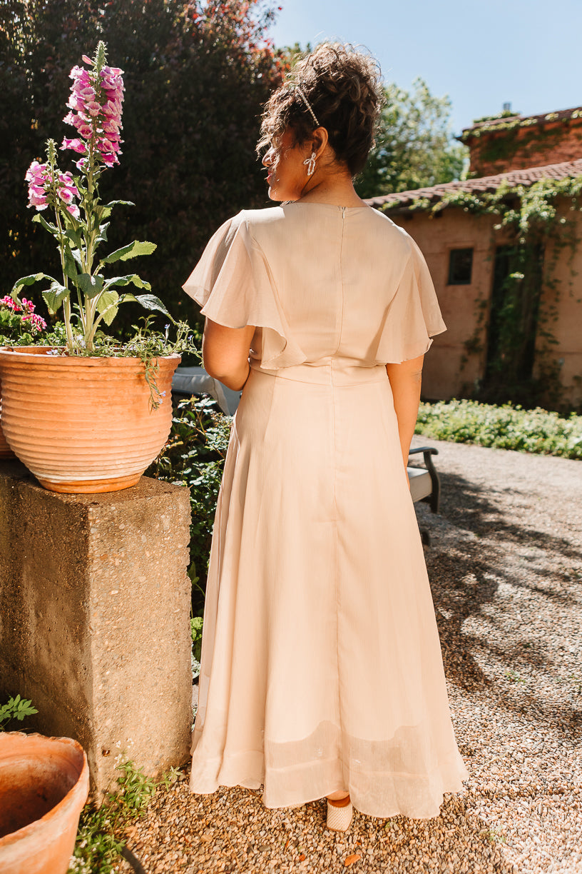 A woman with curly hair stands outdoors, facing away, wearing Ivy City Co’s Callie Dress in Champagne Chiffon with short flutter sleeves. She is beside a potted plant with pink flowers on a sunny gravel path near a rustic building.