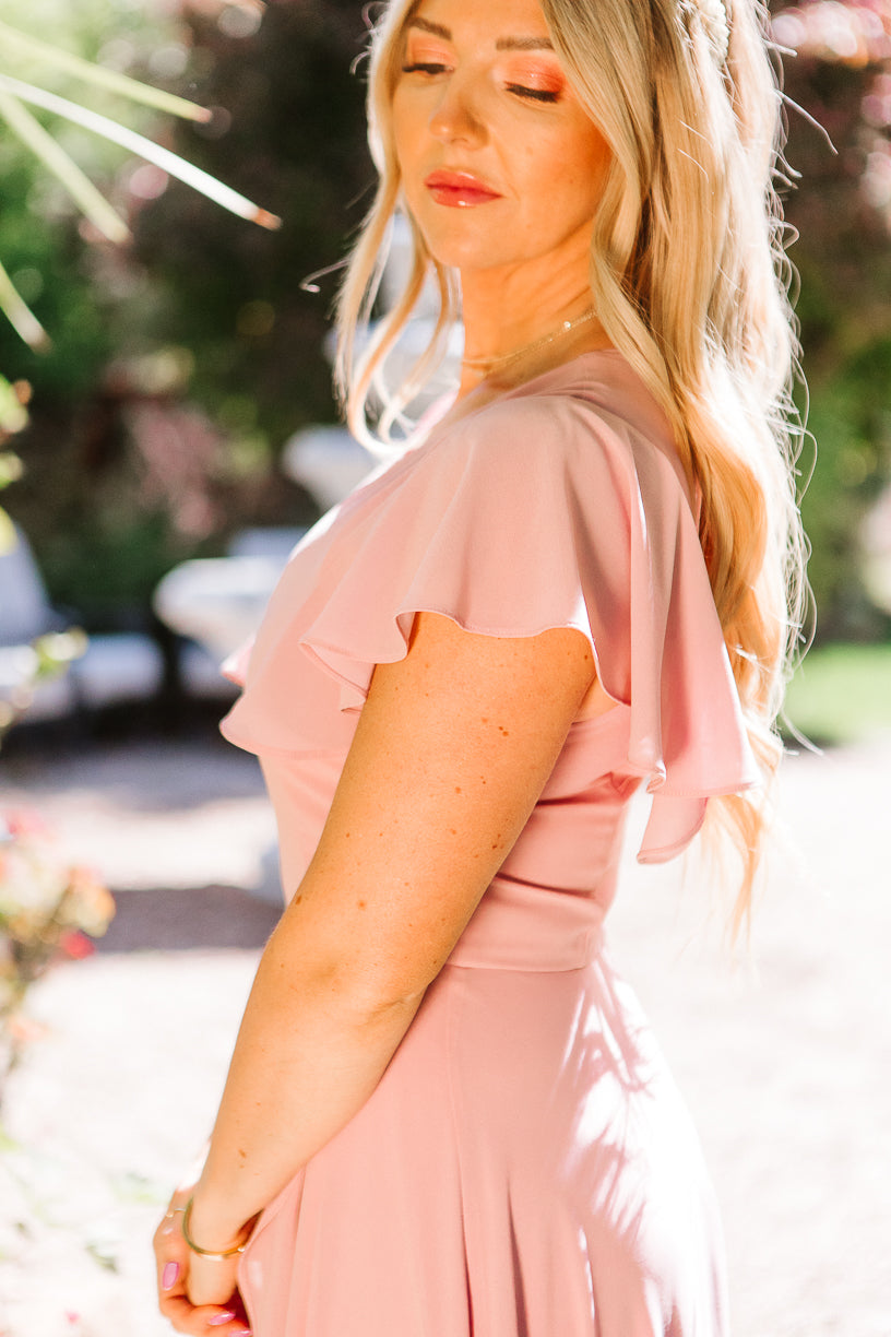 A woman with long blonde hair stands outdoors in sunlight, wearing the Ivy City Co Callie Dress in Blush Chiffon. Eyes closed, she looks peaceful amidst lush greenery and soft natural light.