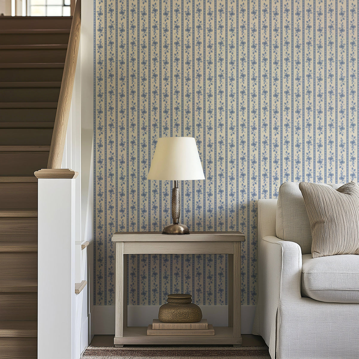 A small table with a lamp and a woven basket beside a sofa sits next to a wooden staircase. The wall is covered with Felicity Floral Wallpaper by Loomwell Home Goods in blue and white, adding charm and elegance to the cozy corner.