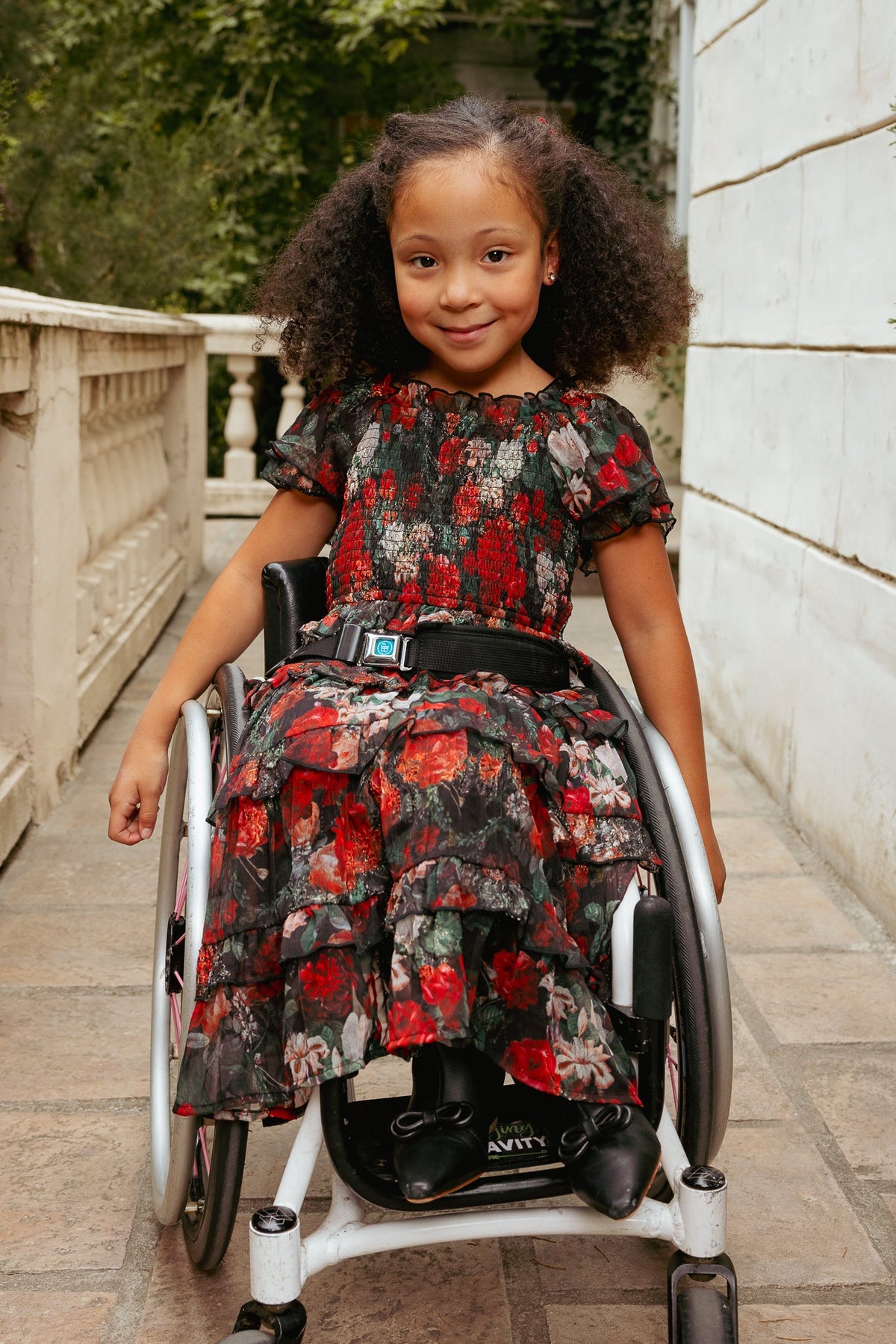 A young girl with curly hair sits smiling in a wheelchair outdoors, next to a white wall and greenery, wearing the Ivy City Co Mini Winnie Dress in Midnight Roses.