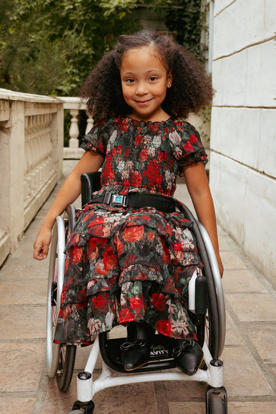 A young girl with curly hair sits smiling in a wheelchair outdoors, next to a white wall and greenery, wearing the Ivy City Co Mini Winnie Dress in Midnight Roses.