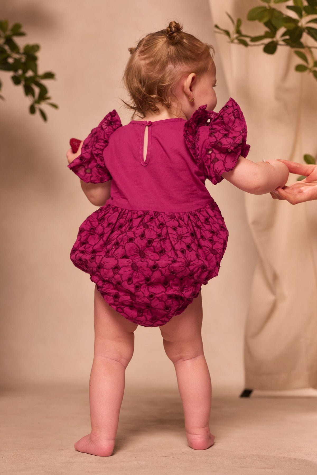 A baby stands barefoot, holding an adult’s hand and facing away from the camera, dressed in the Ivy City Co Baby Hattie Romper in Magenta—a charming embroidered cotton romper with puff sleeves and floral patterns, perfect for an adorable Easter outfit moment.